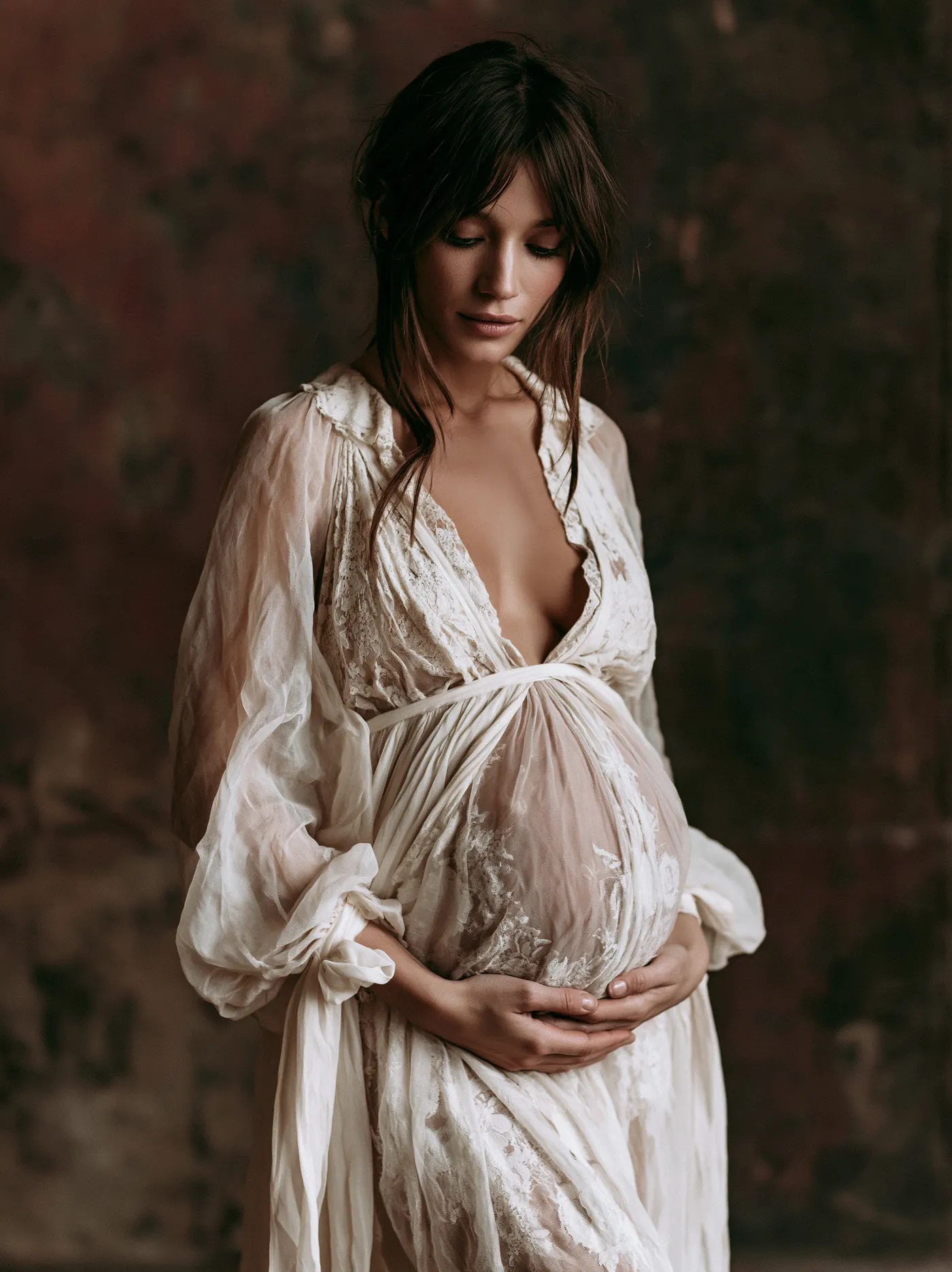 At-home maternity portrait in a pale robe against a dark wall in Los Angeles